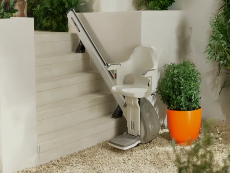 A stair lift is mounted on a set of outdoor stairs leading upward against a light gray or white wall. The residential lift has a white molded seat and footrest, attached to a track set on the stairs. A lush green plant sits in a bright orange pot beside the lift.